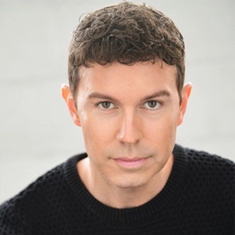 A headshot of Ashley Wells, a man with short, curly brown hair looking directly at the camera. He is wearing a black crew-neck sweater against a neutral, light-colored background.