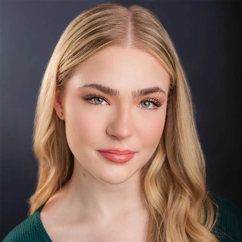 A young woman with long blonde hair and fair skin looks at the camera, smiling gently. She wears natural makeup, with defined eyebrows and pink lipstick. Dressed in a green top, Samme Sultan channels Kitana’s poise against a dark, softly blurred background.