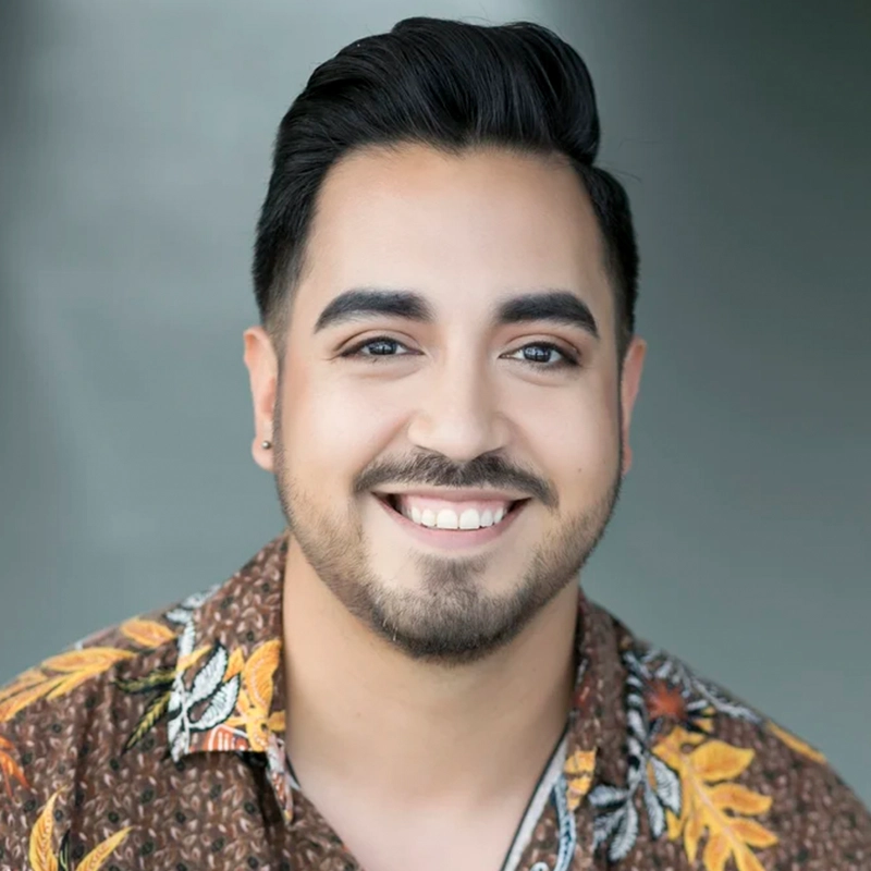 A young man, Joey Rodriguez, with neatly styled dark hair and a trimmed beard smiles warmly at the camera. He wears a brown floral shirt with yellow and orange patterns. The background is plain and blurred, highlighting his friendly expression.