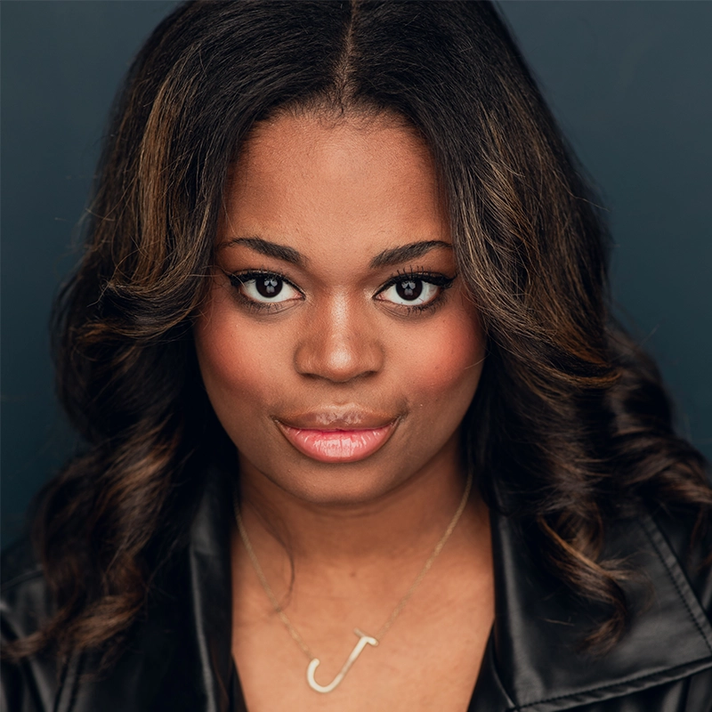 A young woman with medium-dark skin and wavy, shoulder-length hair smiles softly. She wears a black leather jacket and a 