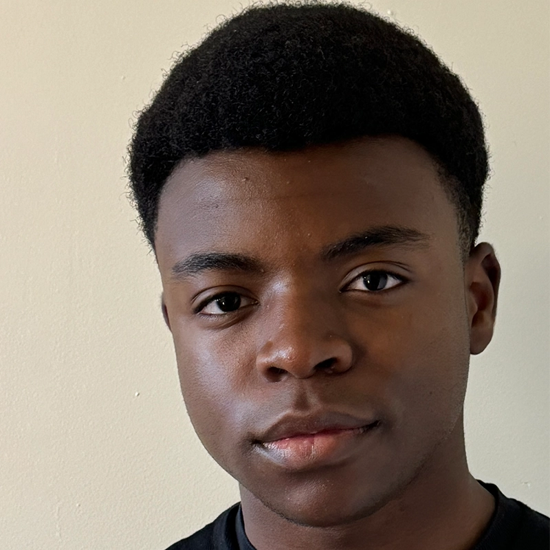 A young Black man with short, natural curly hair looks directly at the camera with a neutral expression. He is wearing a black shirt and standing against a plain light-colored wall, in soft, even lighting—photographed in the style of Anjalee Hardy.