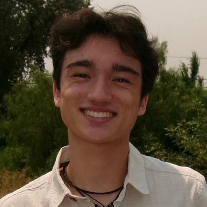 A young man with wavy brown hair and fair skin smiles outdoors. He wears a light-colored collared shirt and two black necklaces. Green trees and bushes fill the blurred background, and the lighting suggests a bright, sunny day.