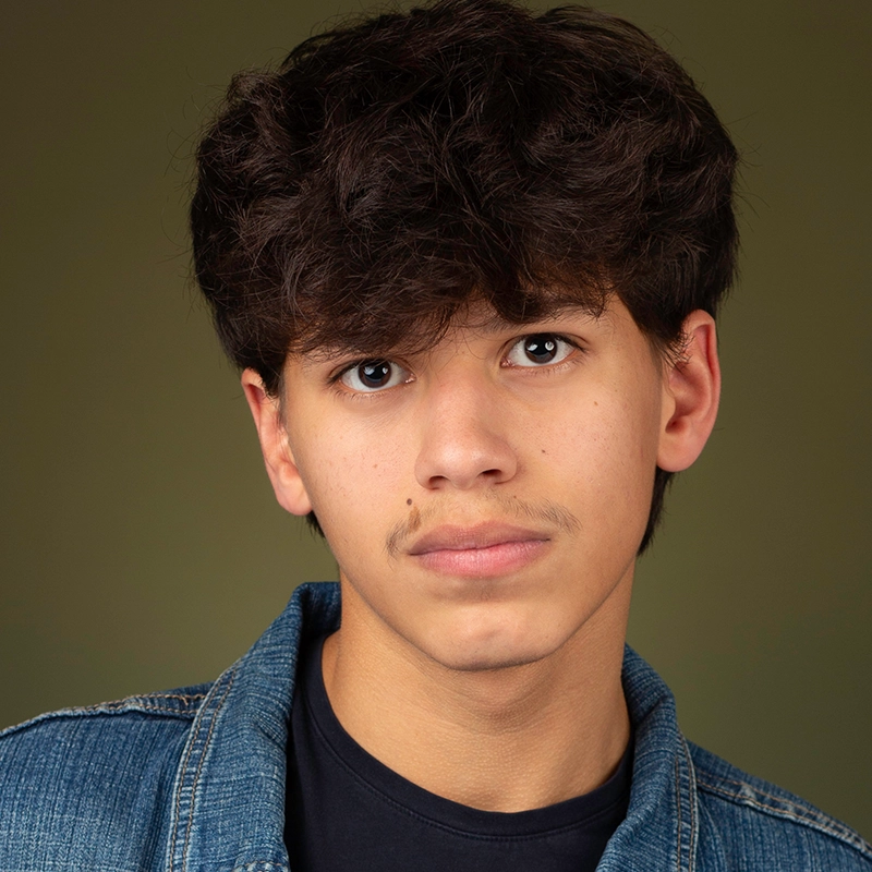 A teenage boy with wavy dark brown hair and light brown skin looks directly at the camera with a neutral expression. He is wearing a blue denim jacket over a black shirt—an image reminiscent of Coral Rosario-Laboy’s portraits. The background is olive green.