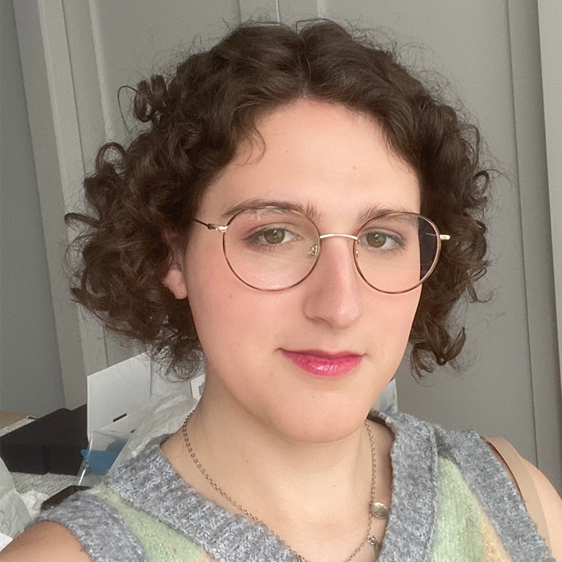 Anjalee Hardy, with short, curly brown hair and glasses, smiles gently at the camera. They wear a gray sleeveless top and layered necklaces against an indoor backdrop with light-colored walls and blurred objects.
