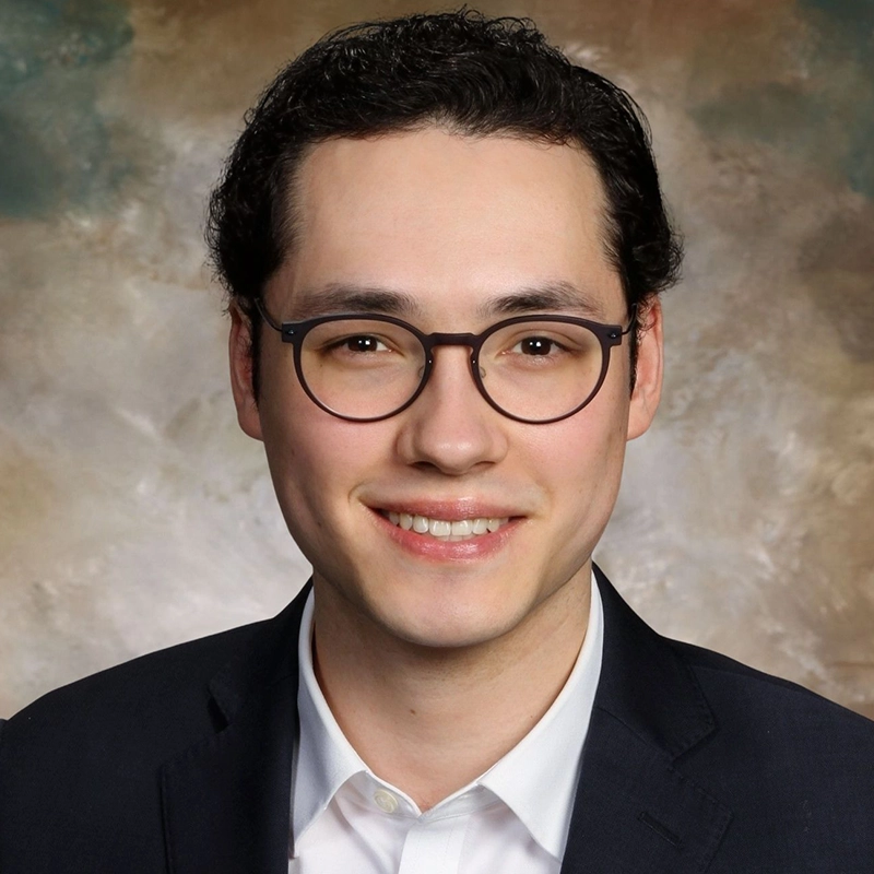 A young man with short, dark curly hair and round glasses smiles at the camera. He is wearing a dark suit jacket and a white collared shirt. The background is a soft, blurred blend of brown and green tones, giving the image a professional portrait look.
