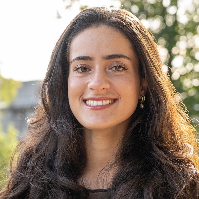 A young woman, Anjalee Hardy, with long, wavy brown hair is smiling outdoors. She is wearing a black top and a small hoop earring with a dangling pearl. Sunlight highlights her hair as green foliage blurs in the background.
