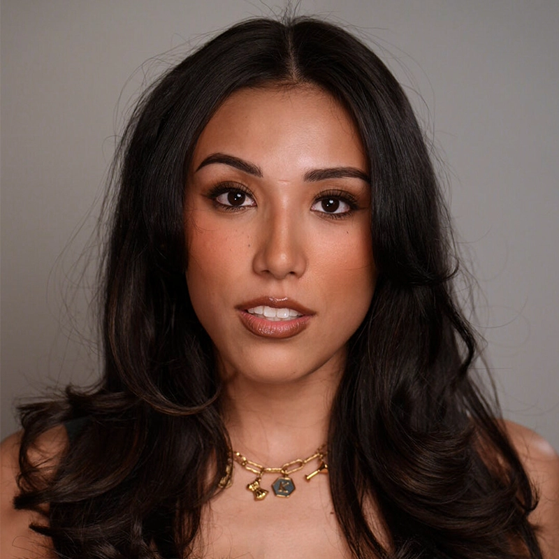 A woman with long, wavy dark hair wears natural makeup with bold brows and nude lipstick. Kayla Hatashita gazes directly at the camera with a neutral expression, wearing a gold necklace and a dark top against a plain, light background.