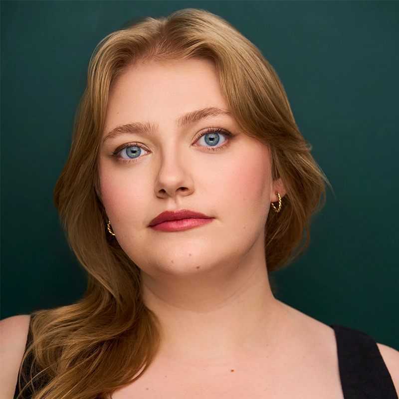 Kayla Hatashita, a young woman with wavy blonde hair and blue eyes, gazes confidently at the camera. She wears light makeup, gold hoop earrings, and a black top against a solid dark green background with soft lighting highlighting her features.