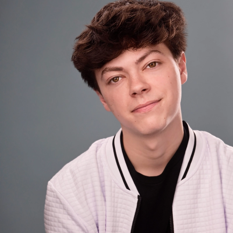 Jaxon Keller, a young person with fair skin and short, wavy brown hair, smiles softly at the camera. They wear a textured white jacket with a black collar and a black shirt underneath against a plain, muted gray background.