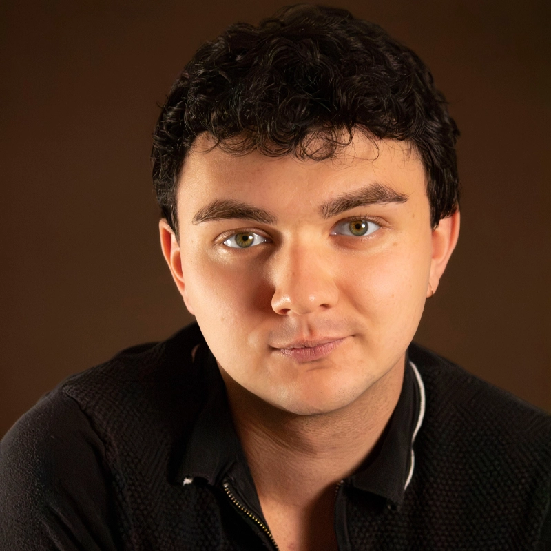 A young man with short, curly dark hair and hazel eyes gazes softly at the camera. He wears a black zip-up top with a textured pattern. The background is solid brown, giving the portrait a warm and neutral tone, highlighting Ryan P. Kennedy’s thoughtful expression.