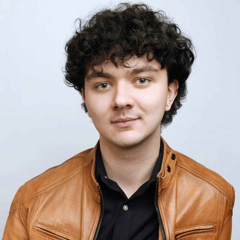 A young person with curly dark hair, light skin, and green eyes faces the camera, slightly smiling. Ryan P. Kennedy wears a brown leather jacket over a black collared shirt against a plain, light-colored background.