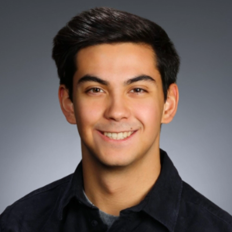 A young man with short, dark hair smiles at the camera. He is wearing a dark collared shirt over a gray undershirt. The background is a smooth gradient from dark gray at the top to lighter gray at the bottom.