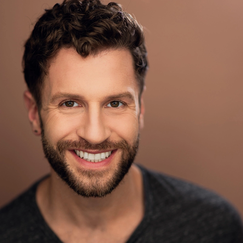 A man with curly brown hair, a trimmed beard, and bright white teeth smiles warmly at the camera. He is wearing a dark V-neck shirt and stands against a plain, softly lit brown background. The overall mood is friendly and approachable.