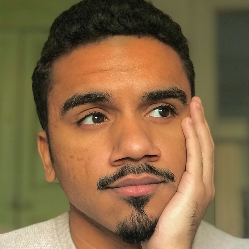 Kyle Castillo, a young man with short, curly black hair and a goatee, rests his head on his hand, gazing thoughtfully into the distance. He has warm brown skin, expressive eyebrows, and is wearing a light-colored top against a softly blurred background.