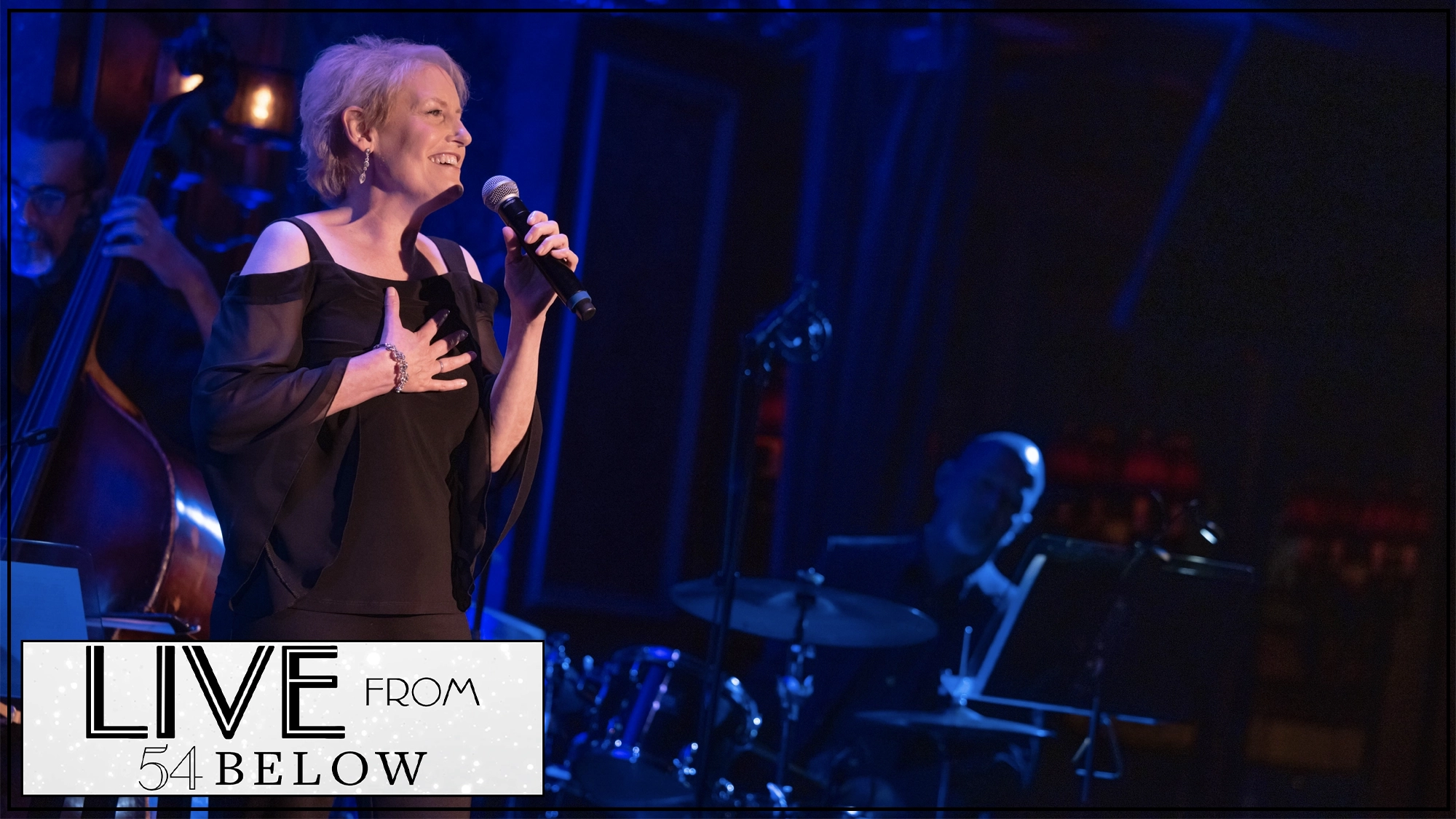 A woman with short blond hair sings into a microphone on stage, wearing a black dress with sheer sleeves. Performing live from 54 Below, Liz Callaway delivers a heartfelt Sondheim number as musicians play behind her.