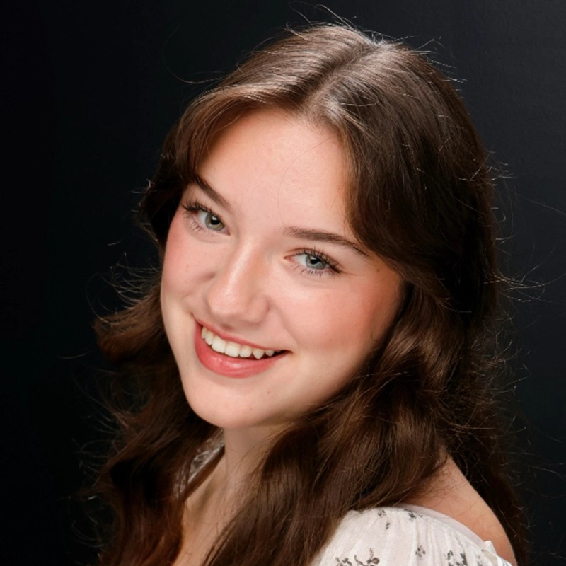 A young woman with long, wavy brown hair and blue eyes smiles at the camera. Reminiscent of Kitana, she wears a white top with a subtle pattern. The black background and soft lighting make her natural features stand out brilliantly.