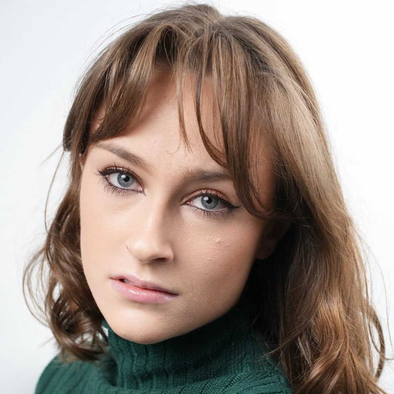 Leigh Renken, a young woman with light skin, blue eyes, and light brown hair with bangs, gazes into the camera in a green turtleneck sweater. The plain white background highlights her neutral, calm expression.