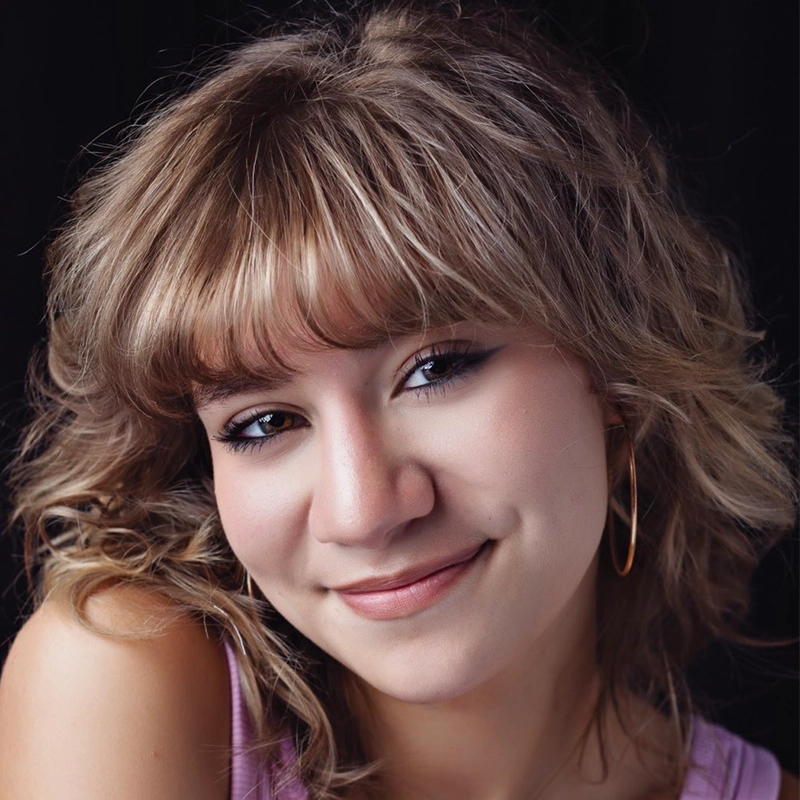 Quinton Robinson, a young person with wavy, light brown hair and bangs, smiles gently at the camera. They wear hoop earrings and a sleeveless lavender top against a black background, highlighting their relaxed, friendly expression and natural makeup.