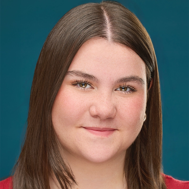 A young woman with straight brown hair and fair skin smiles slightly. She is wearing a red top, posed against a solid teal background—a portrait styled by Quinton Robinson. Her hair is parted in the middle, and she looks directly at the camera.