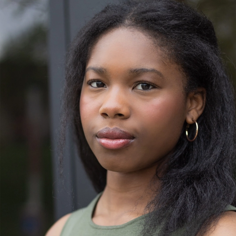 A young Black woman with straight, shoulder-length hair wearing a sleeveless green top and small gold hoop earrings stands outdoors, looking directly at the camera with a neutral expression. The background is softly blurred.