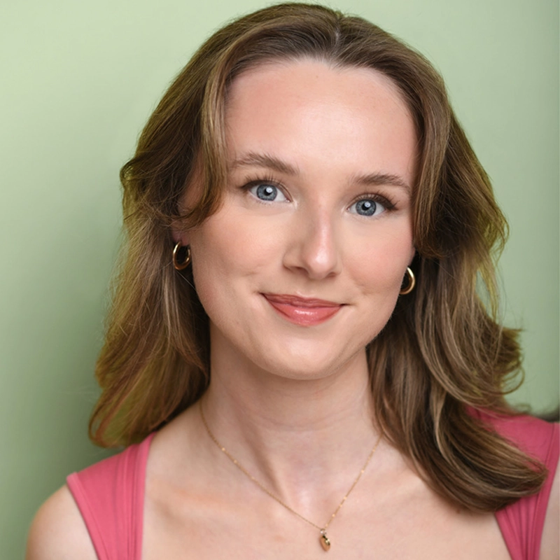 Madison Keenoy, a woman with light skin, wavy brown hair, and blue eyes, smiles gently. She wears gold hoop earrings, a delicate gold necklace, and a sleeveless pink top against a soft green background with bright, even lighting.
