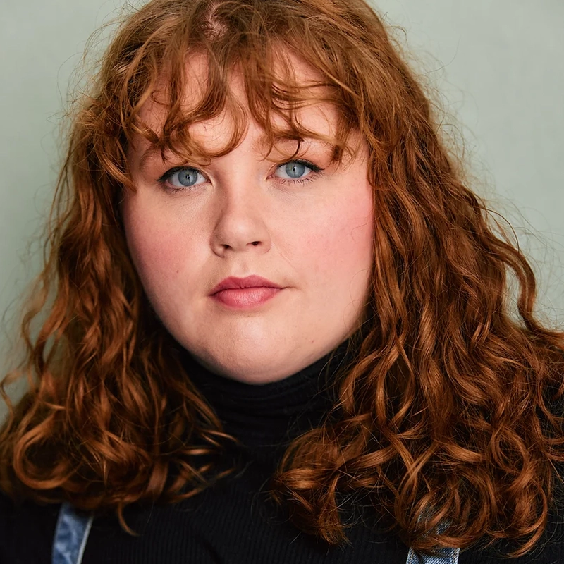 Madison Keenoy, with curly, shoulder-length red hair and blue eyes, looks directly at the camera. They wear a black turtleneck sweater and denim overalls against a plain, light-colored background for a simple, neutral setting.