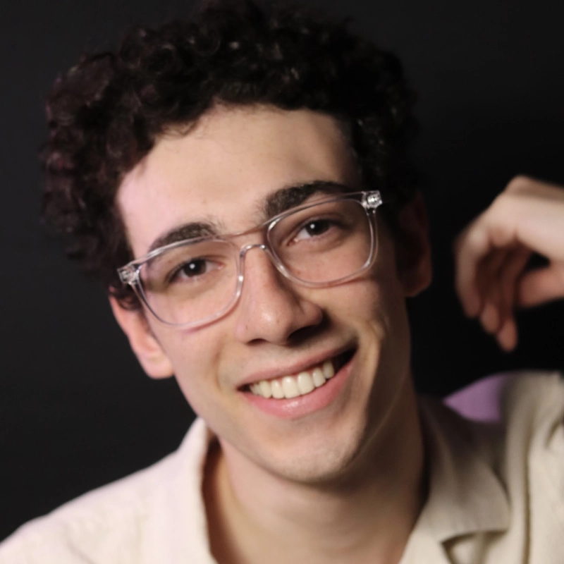 A young person with short, curly dark hair and clear glasses smiles at the camera. Lucas Marinetto wears a light-colored shirt and is posed in front of a dark background, with one arm bent and a relaxed, friendly expression.