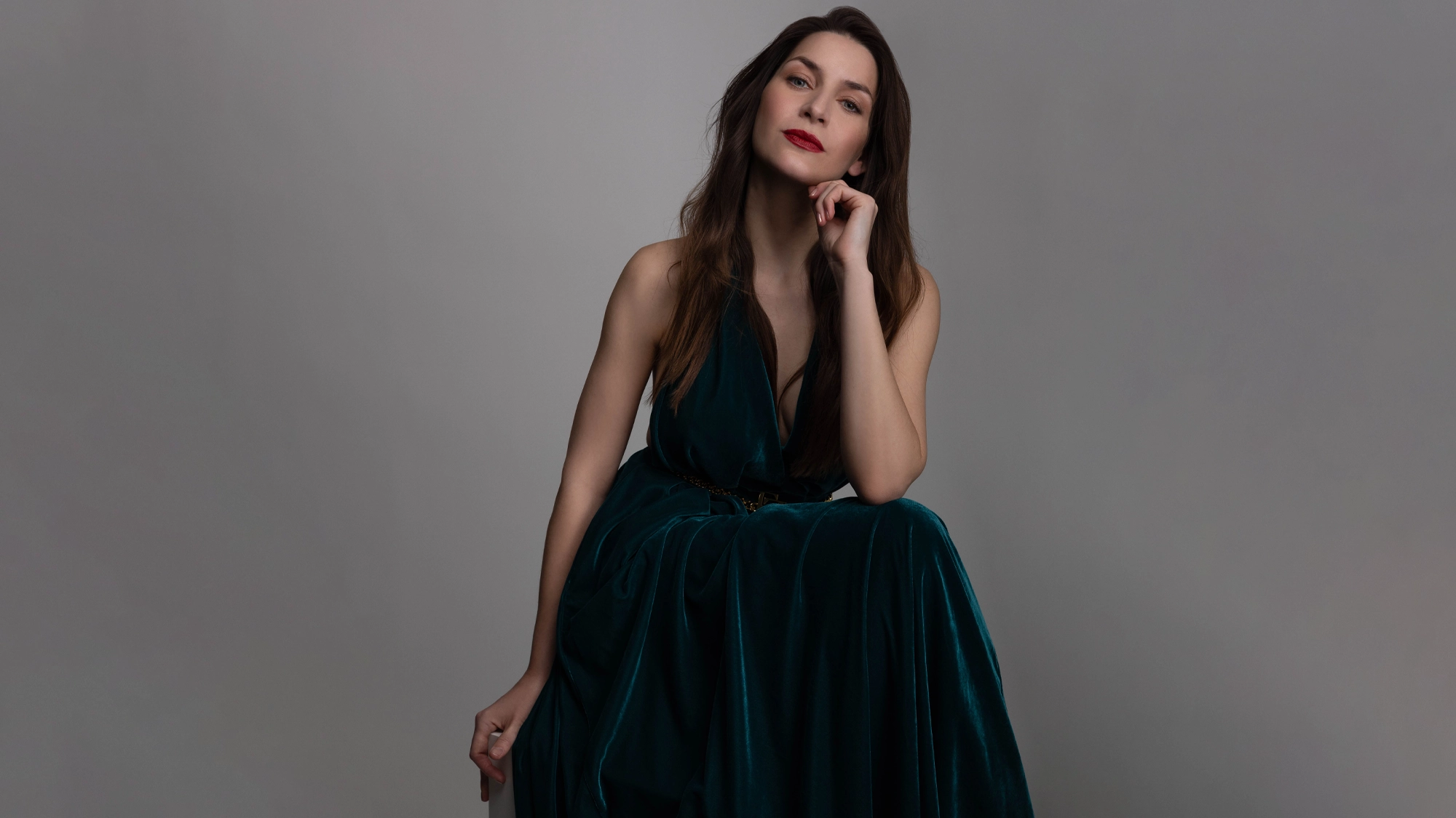Martina Barta, in a sleeveless dark teal velvet dress, sits on a stool against a plain gray background. She rests her chin on her hand, looking confidently at the camera—perfectly poised for a Burt Bacharach Tribute or 