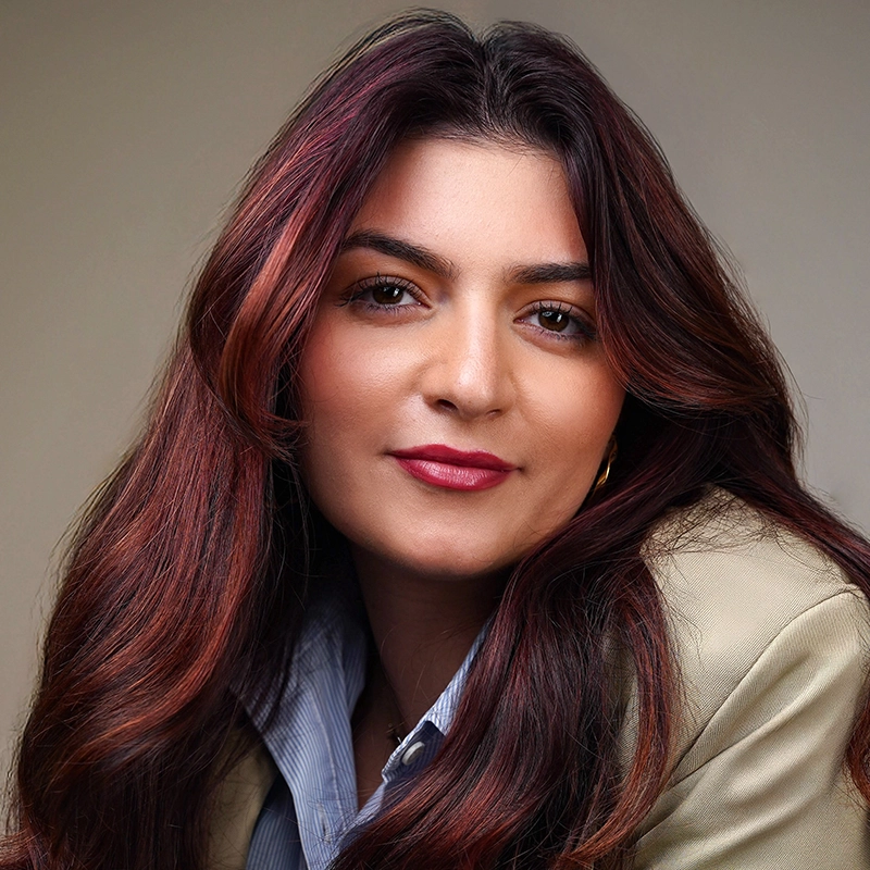 Grace Whetstone, with long, wavy dark brown hair and a soft smile, looks at the camera. She wears a beige blazer over a blue collared shirt, her makeup natural with bold brows and red lipstick against a plain, neutral background.