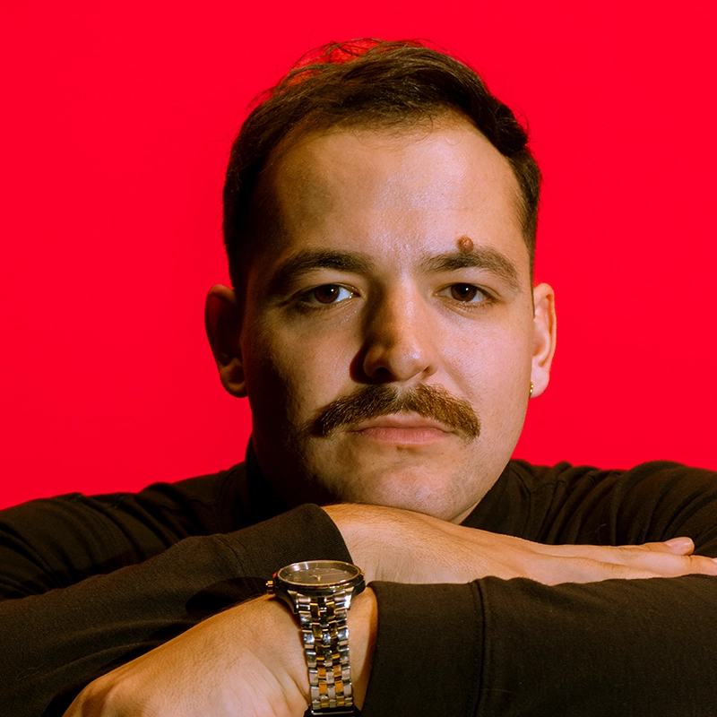 Max Marsillo, a man with short dark hair, a thick mustache, and a large mole above his left eyebrow, rests his arms on a surface. He wears a black shirt and a silver wristwatch against a solid, bright red background, his expression neutral.