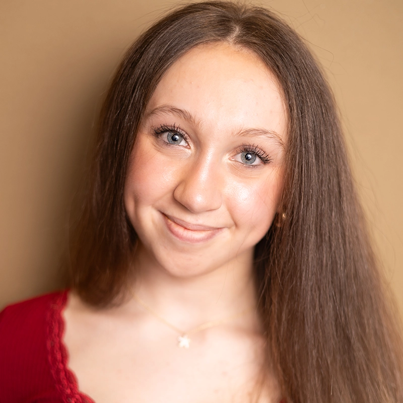 Maya Annunziato, a young woman with long straight brown hair and blue eyes, smiles softly at the camera. She wears a red top with lace trim and a delicate gold necklace with a small star pendant against a warm beige background.