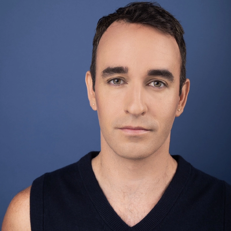A person with short dark hair and light skin looks calmly at the camera. Wearing a sleeveless, dark V-neck top and small earrings, they stand against a solid blue background. Their neutral expression gives a composed, Matthew Meade-inspired presence.