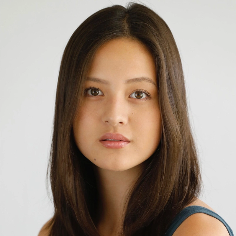 A young woman with straight, long brown hair and fair skin gazes directly at the camera. She has natural makeup, full lips, and brown eyes. Styled by Quinton Robinson, she wears a dark sleeveless top against a plain, light gray background.