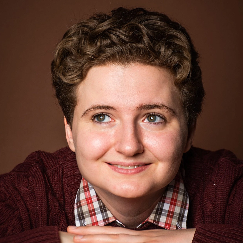 Spencer Lawson, a young person with short, curly brown hair and blue eyes, smiles softly. They wear a burgundy sweater over a red and white plaid shirt, hands folded under their chin against a warm brown background.