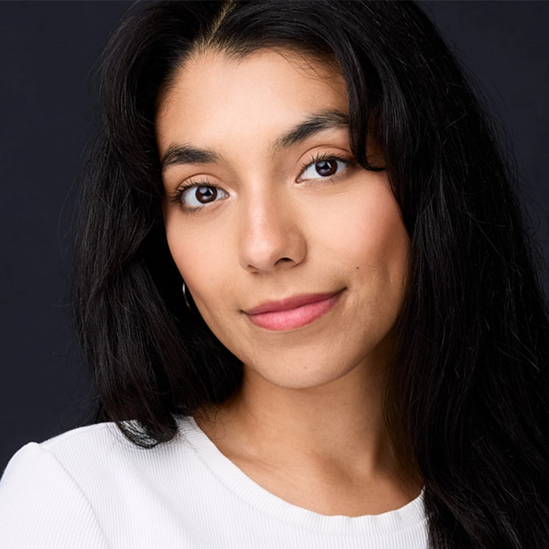 Isabel Gonzalez, a young woman with long, wavy black hair and medium skin tone, smiles gently. She has brown eyes, subtle makeup, and wears a white ribbed top. The plain dark background highlights her confident gaze at the camera.