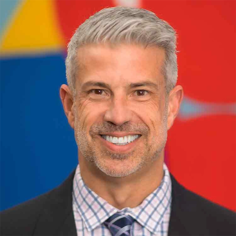 A professional portrait of Ashley Wells, a middle-aged man with short, gray hair and a light beard, smiling at the camera. He is wearing a dark blazer over a light-colored, checkered button-down shirt and a patterned tie.