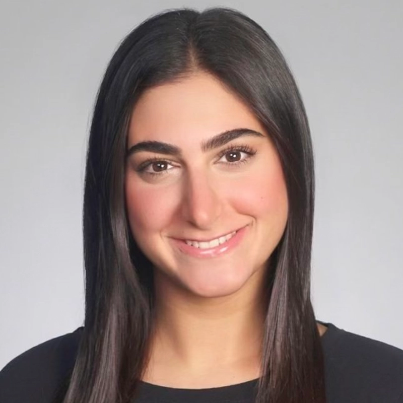 A woman with straight, dark brown hair and fair skin smiles softly. She is wearing a black top and has well-groomed eyebrows. The plain light gray background gives this Kitana headshot a professional appearance.