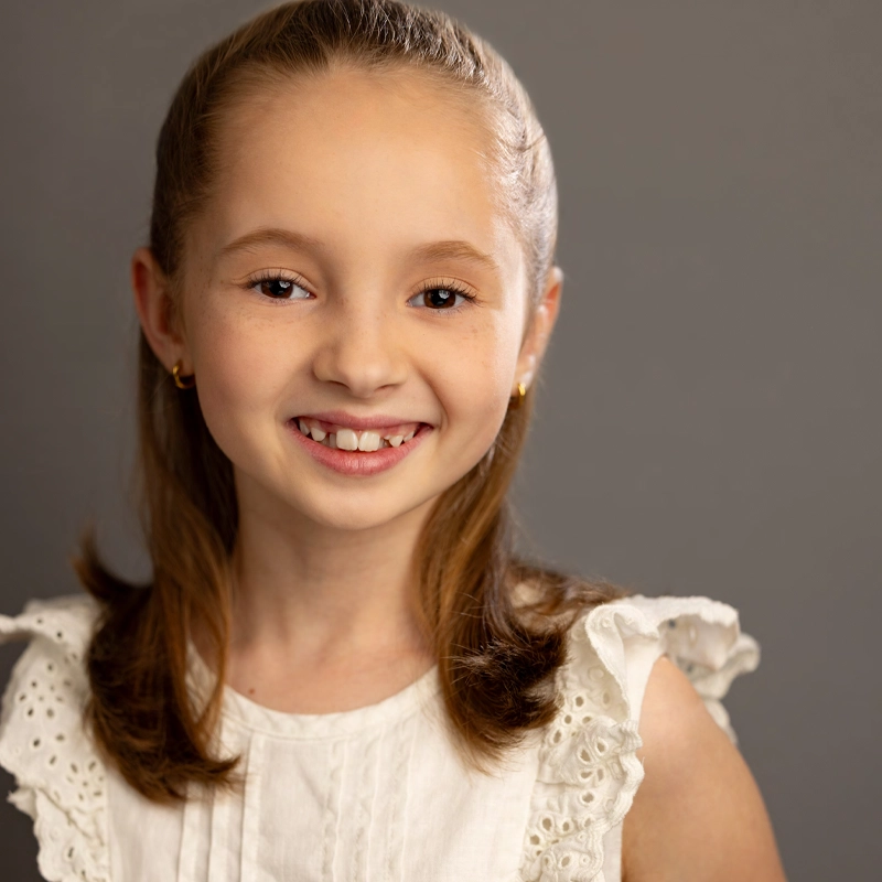 A young girl with light skin and brown hair smiles at the camera. Styled by Robin Miller, she wears a white eyelet dress with ruffled sleeves and small gold hoops. Her hair is neatly pulled back, and she stands before a plain, gray background.