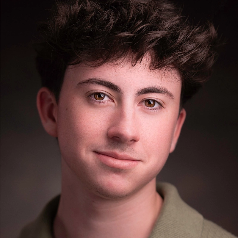 Quinton Robinson, a young person with light skin and brown, wavy hair, looks at the camera with a gentle smile. Wearing a green collared shirt, Quinton's brown eyes stand out against the softly blurred, dark neutral background.