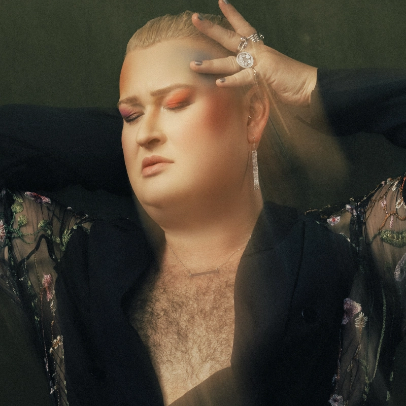 Ryan James Monroe, with long blond hair, bold pink eyeshadow, and a sheer floral top, poses with eyes closed and arms raised. Rings, earrings, and a necklace adorn them as chest hair shows beneath an expression of calm introspection.