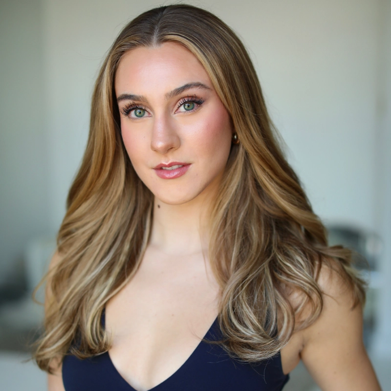 A young woman with long, wavy blonde hair and light green eyes looks confidently at the camera. She wears natural makeup and a navy blue sleeveless top with a V-neck. The background is softly blurred in neutral tones.