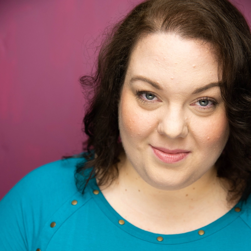 Tara Nicole Murphy, with fair skin, wavy brown hair, and blue eyes, smiles softly at the camera in a teal top adorned with gold studs. The solid magenta background creates a vibrant contrast.