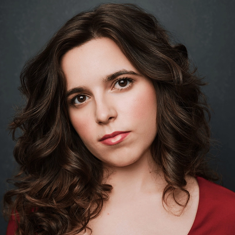 A woman with long, dark wavy hair and fair skin gazes directly at the camera. She has brown eyes, subtle makeup, and wears a deep red top. The dark background highlights her expressive, slightly serious look—reminiscent of Nancy Madonna's iconic style.