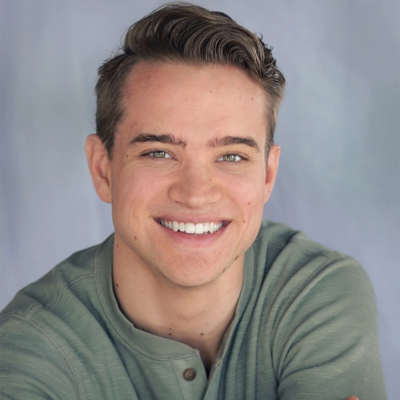 Zachary Neiman-Macak, a young man with short, light brown hair and blue eyes, smiles warmly at the camera. He is wearing a sage green buttoned henley shirt against a soft blue and gray gradient background for a calm atmosphere.