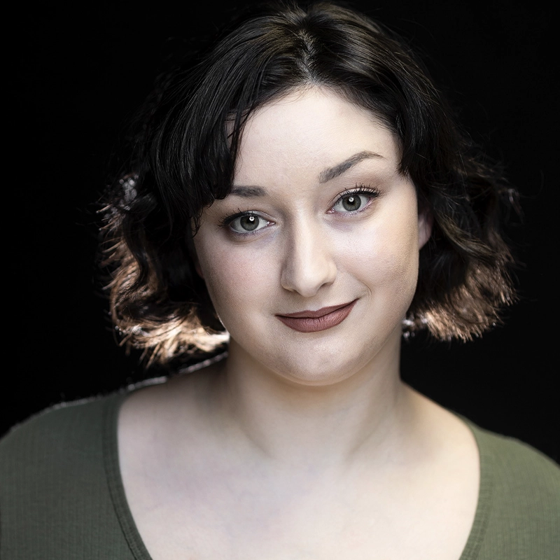 A woman with fair skin and short, wavy dark hair is softly smiling. She wears a muted green top and natural makeup. The background is dark, highlighting her illuminated face and shoulders. Her expression is calm and confident.