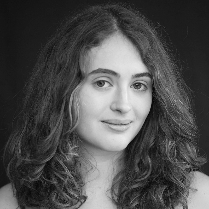 A young woman with long, wavy hair and light skin looks at the camera with a gentle smile. Anjalee Hardy stands in front of a dark background, wearing a light top. The black and white photo features soft lighting that highlights her features.