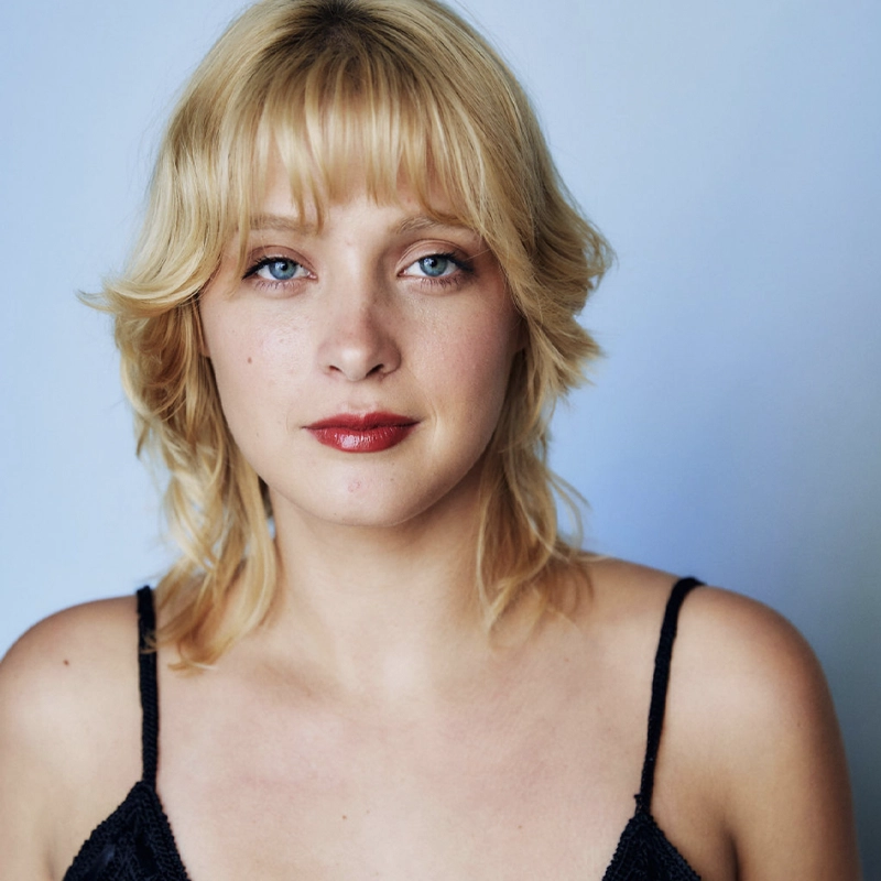 A young woman with short, wavy blonde hair and blue eyes looks directly at the camera. Reminiscent of Madison Osment, she wears a black spaghetti strap top and red lipstick against a soft light blue background.