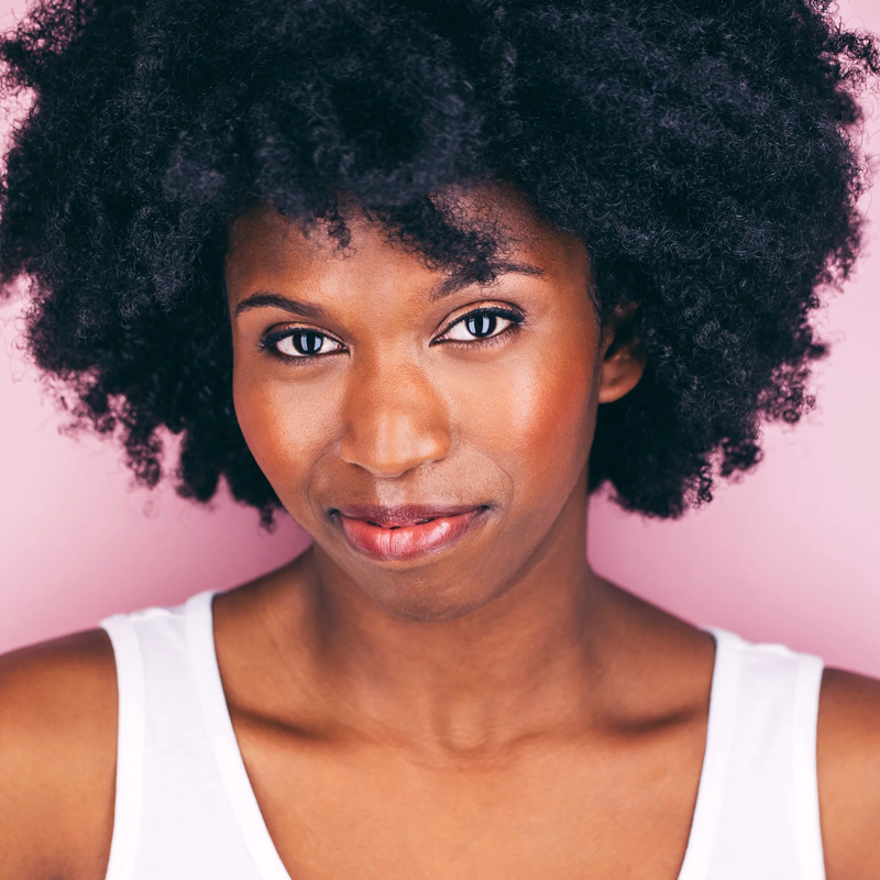 Rachel Pegram, with dark skin and natural, voluminous curly hair, poses confidently against a light pink background in a white tank top. She smiles gently at the camera under soft, flattering lighting.