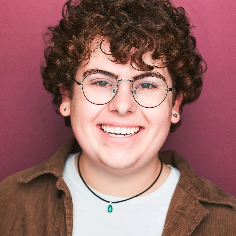 Ethan Poisson, a young person with short, curly brown hair and glasses, smiles broadly against a pink background. They wear a brown corduroy shirt over a white top and a black necklace with a green pendant, plus earrings in both ears.