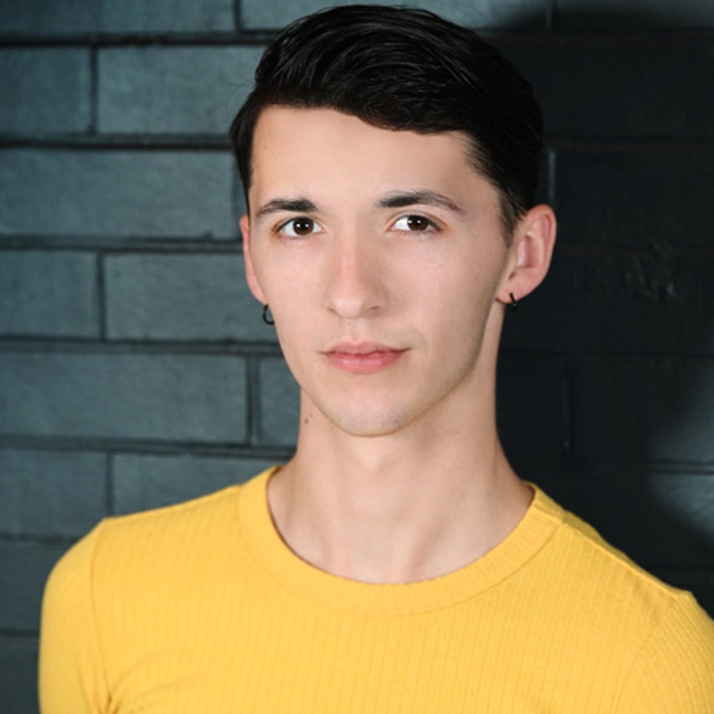A young person with short dark hair, light skin, and a small hoop earring poses in front of a dark brick wall. Wearing a fitted yellow shirt, Kaleb Purswell looks directly at the camera with a neutral expression.